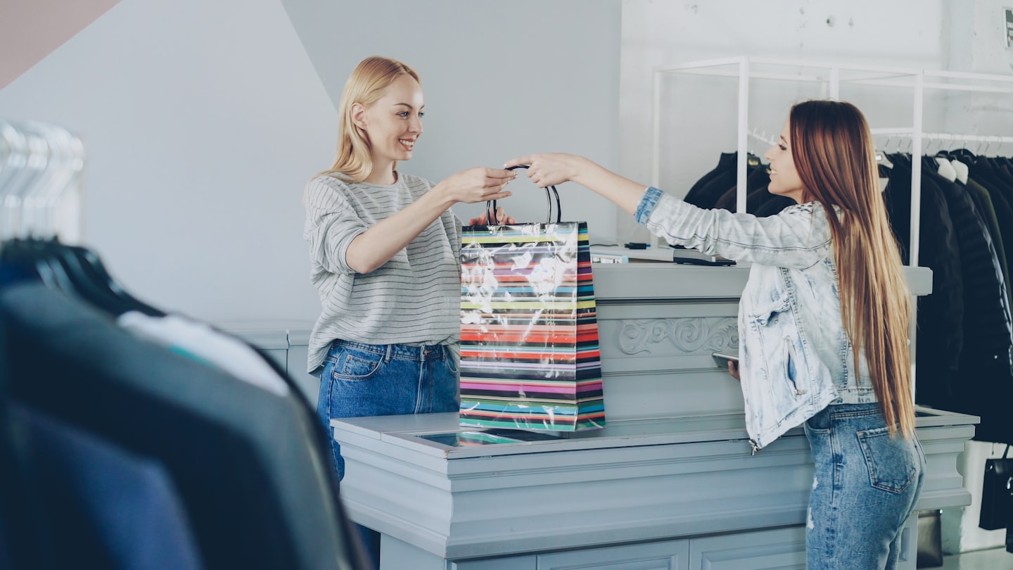 Shop assistant hands customer a colorful shopping bag at a clothing store.