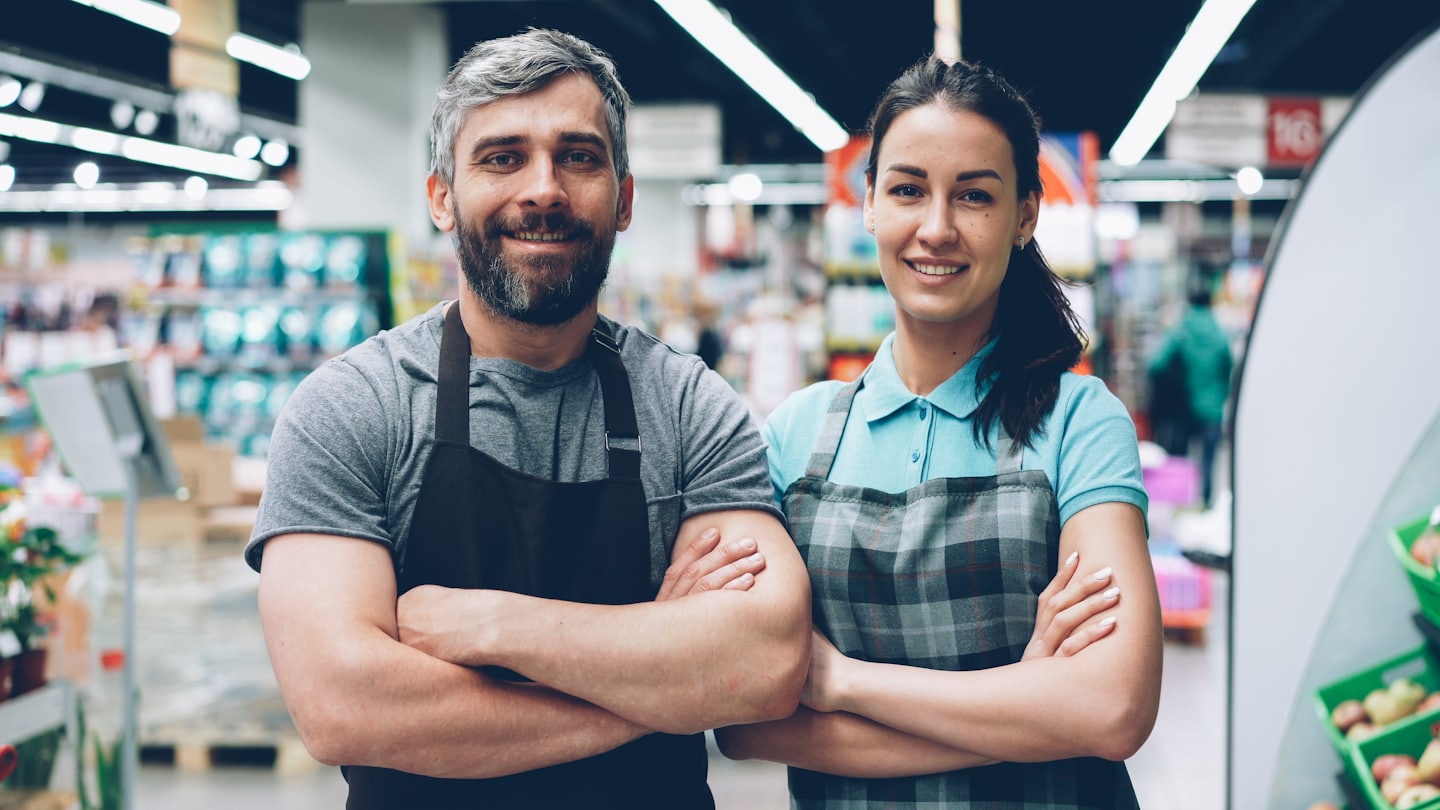 Two smiling store employees in aprons stand in a supermarket aisle.
